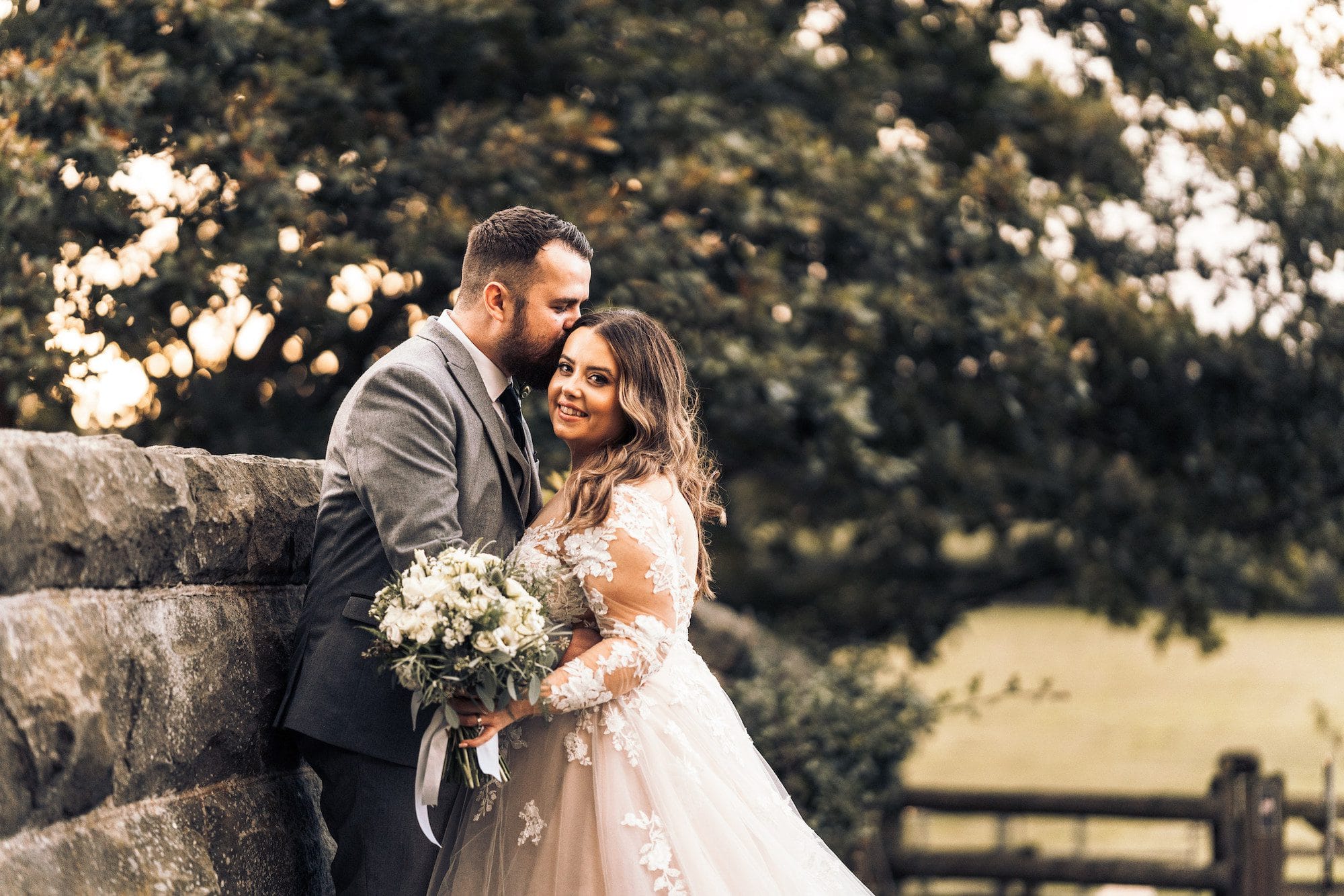 Newlywed couple embrace, leaning against the stone wall of the old railway bridge at Tower Hill Barns. Green trees and rolling fields can be seen in the background.