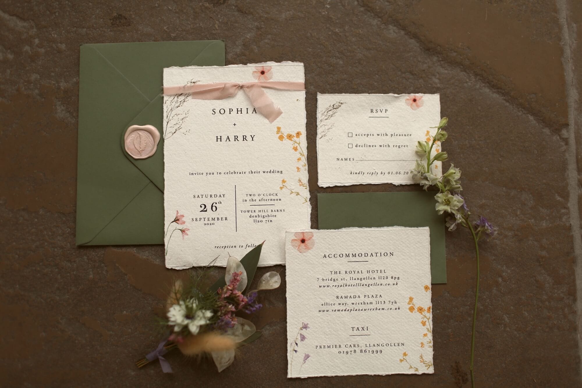 A flat lay image of beautiful wedding invitations. The invitations are printed on off-white textured paper with floral accents, while the envelopes are a forest green colour with a gold seal. The invitations are British-sourced and eco-friendly.
