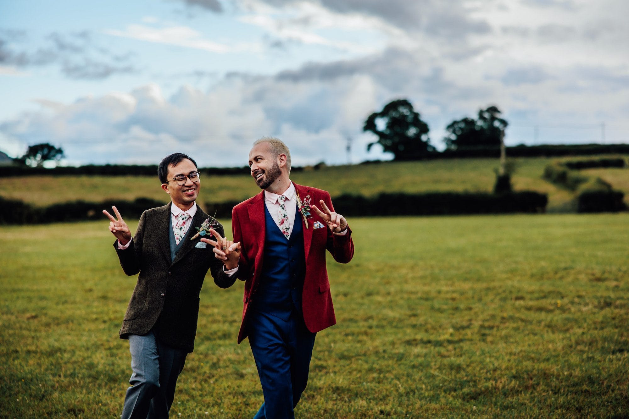Two newlywed grooms skip happily through a field, doing peace signs following their same-sex wedding. In the background rolling views of the North Wales countryside and a blue sky.
