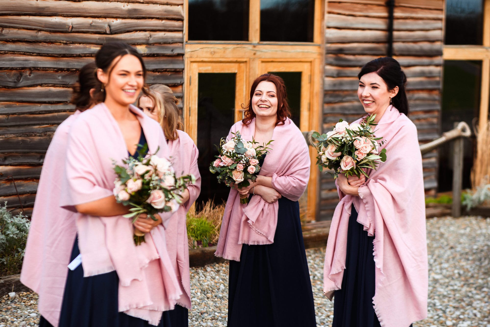 Bridesmaids assemble outside Tower Hill Barns dressed in floor length navy gowns with blush pink pashminas wrapped tightly around their shoulders to keep out the February chills.
