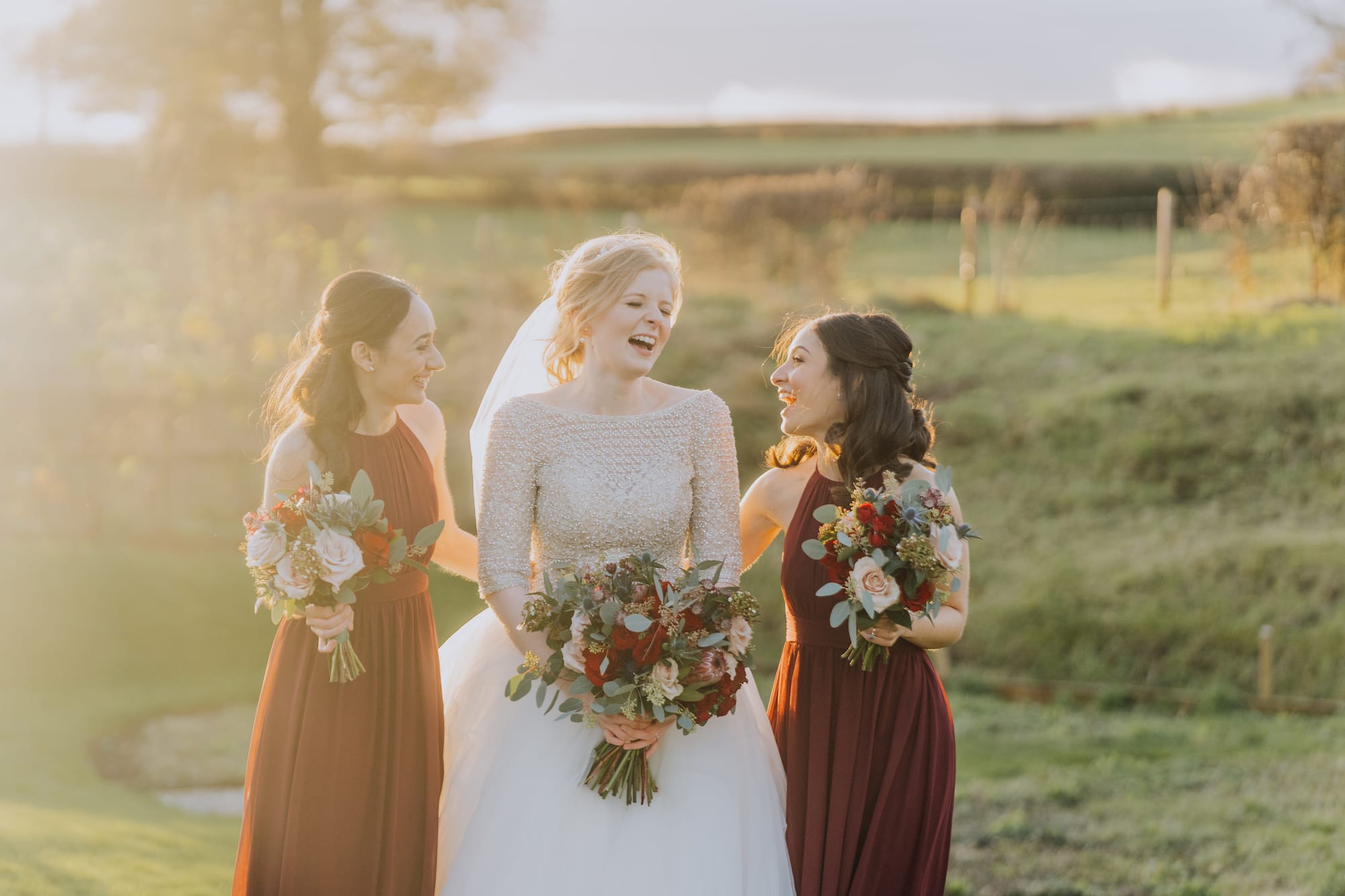 bride in a white sparkly dress and two bridesmaids in rich deep red toned dresses basking in the golden sunshine in winter Tower Hill Barns